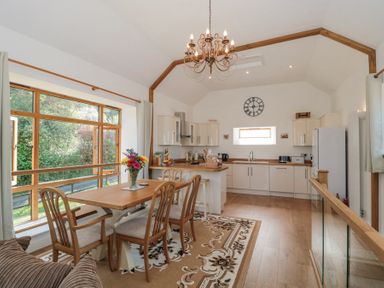 A kitchen and dining area with wooden table, chairs, chandelier, large window, and kitchen cabinets at Newlands Barn in Wellington