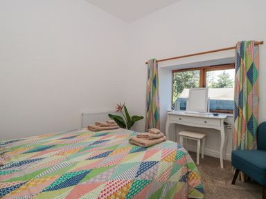 A bedroom with two beds covered in colorful patchwork quilts a white dressing table with a mirror and stool near a window with patterned curtains and a blue chair at Newlands Barn in Wellington