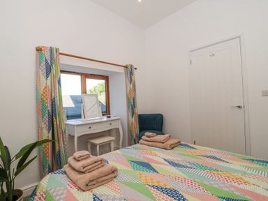 A bedroom with a bed covered by a patterned quilt and folded towels a white dressing table under a window with patterned curtains a green chair and a potted plant at Newlands Barn in Wellington