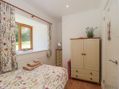 A bedroom with a bed covered with floral bedding a wardrobe and chest of drawers next to a window with floral curtains at Newlands Barn in Wellington
