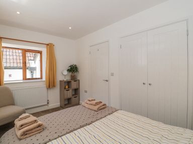 A bedroom with a bed, folded towels on the bed, a window with yellow curtains, a small cabinet with a plant and mirror, and white doors at Newlands Barn in Wellington
