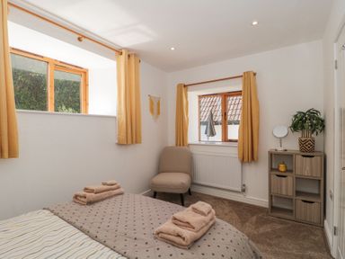 A bedroom with two windows yellow curtains a beige chair a small shelving unit with a plant and mirror and a bed with folded towels at Newlands Barn in Wellington