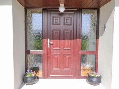 A wooden front door with textured glass panels on either side and flower pots in black containers at Teach Ards in Kilgarvan County Kerry
