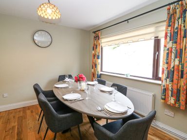 A dining room with a wooden oval table set with plates and glasses six chairs colorful curtains and a wall clock at Teach Ards in Kilgarvan County Kerry