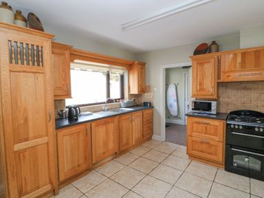 A kitchen with wooden cabinets a black stove a microwave kettle and an ironing board in an adjoining room at Teach Ards in Kilgarvan County Kerry