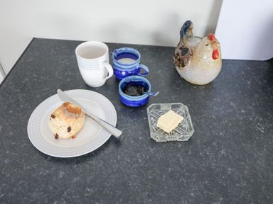 A kitchen counter with a scone on a plate with a knife a white mug two small blue pots a glass dish of butter and a ceramic chicken at Teach Ards Kilgarvan County Kerry