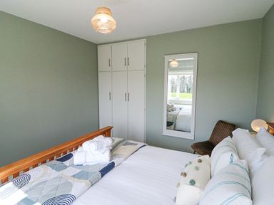 A bedroom with a wooden bed frame white bedding a decorative pillow a chair a wall mirror and white cupboards at Teach Ards in Kilgarvan County Kerry