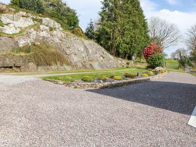 A gravel driveway beside a rock wall with a garden bed and trees at Teach Ards in Kilgarvan County Kerry