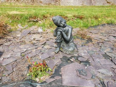 A small statue of a person sitting with head resting on hands on a ground covered with broken slate pieces and a patch of grass in the background at Teach Ards in Kilgarvan County Kerry