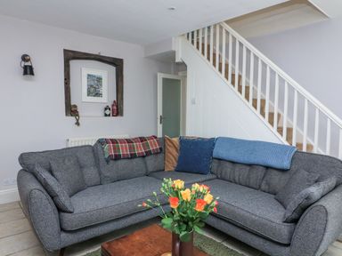 A living room with a grey sofa, throws and cushions, a wooden coffee table with flowers, stairs and a recessed wall shelf at Joymead in Malborough