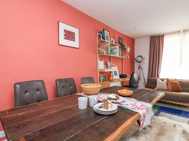 A dining room with a wooden table with food and mugs and chairs against a coral wall with bookshelves and a sofa at Joymead in Malborough