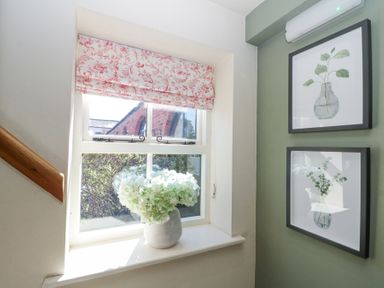 A window with a floral roman blind and a vase with white flowers on the sill next to two framed botanical prints on a green wall at Beaver Cottage in Little Thirkleby near Thirsk