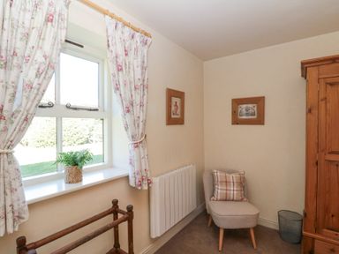 A small room with a window with floral curtains a wooden towel rack a potted plant on the windowsill a small upholstered chair with a pillow two framed pictures on the wall a wooden wardrobe and a waste bin at Beaver Cottage in Little Thirkleby near Thirsk