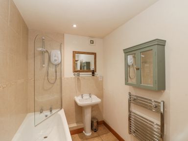 A bathroom with a bathtub shower combination a pedestal sink a wall-mounted mirror a green cabinet and a chrome towel radiator at Beaver Cottage in Little Thirkleby near Thirsk