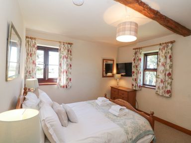 A bedroom with a double bed two windows floral curtains a wooden dresser and a wall-mounted TV at Beaver Cottage in Little Thirkleby near Thirsk