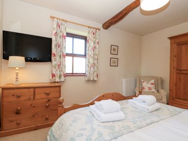 A bedroom with a wooden bed and folded towels a wooden dresser with a lamp a wall-mounted TV floral curtains an armchair and a wooden wardrobe at Beaver Cottage in Little Thirkleby near Thirsk