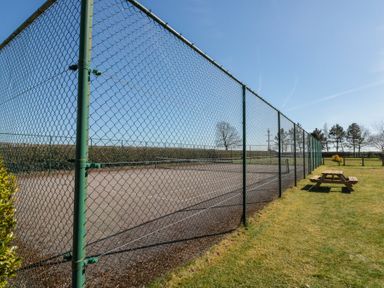 An outdoor tennis court with a green chain link fence and a wooden picnic table on grass at Beaver Cottage in Little Thirkleby near Thirsk