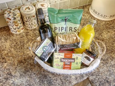 A kitchen countertop with a basket containing wine bottle biscuits crisps tea butter jam and orange juice at Beaver Cottage in Little Thirkleby near Thirsk