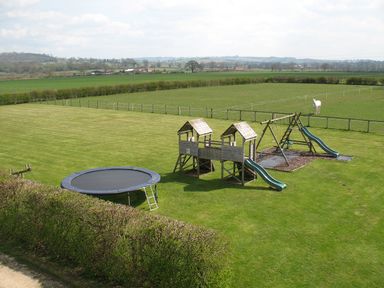 An outdoor playground with a trampoline, wooden play structure with slides, swings, and a field with a white horse at Beaver Cottage in Little Thirkleby near Thirsk