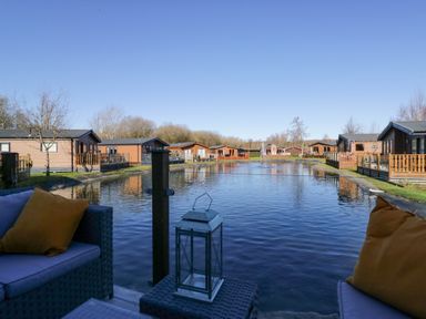 An outdoor seating area with yellow pillows overlooking a pond surrounded by wooden cabins at Coniston View Lodge in Warton near Carnforth