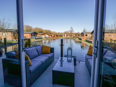 A patio with outdoor sofas and a table overlooking a water canal with lodges on both sides at Coniston View Lodge in Warton near Carnforth