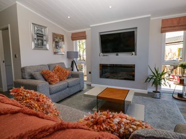 A living room with grey sofas and orange cushions a coffee table a wall-mounted television and a fireplace at Coniston View Lodge in Warton near Carnforth