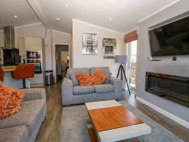 A living room with grey sofas and orange cushions a flat screen tv on the wall and a kitchen area with bar stools at Coniston View Lodge in Warton near Carnforth