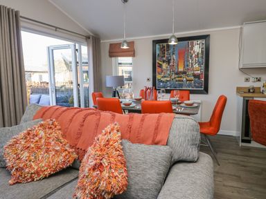 A living room with grey sofa orange cushions and blanket dining table with orange chairs and a painting on the wall at Coniston View Lodge in Warton near Carnforth