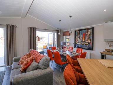 An open living and dining area with a gray sofa and orange cushions a dining table with orange chairs and place settings a painting on the wall and outdoor seating visible through open doors at Coniston View Lodge in Warton near Carnforth