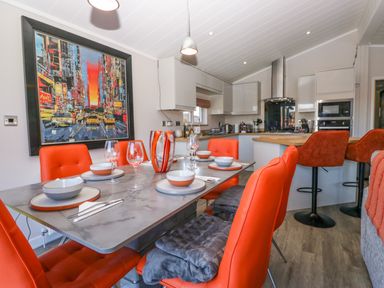 A dining area with a table set for six and orange chairs next to a kitchen with white cabinets and bar stools at Coniston View Lodge in Warton near Carnforth