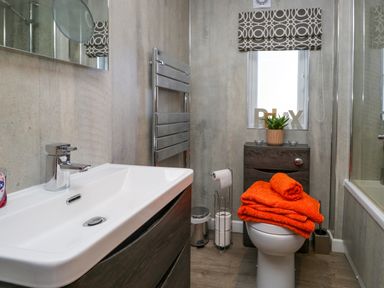 A bathroom with a sink, toilet with orange towels, a small cabinet, a window with patterned blind and a bathtub at Coniston View Lodge Warton near Carnforth