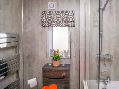 A bathroom with a shower, bathtub, wooden cabinet with a plant and decor, a towel rack, and a window with a patterned blind at Coniston View Lodge in Warton near Carnforth