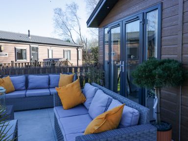 An outdoor seating area with grey cushioned sofas and yellow pillows outside a wooden building with glass doors at Coniston View Lodge in Warton near Carnforth