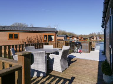 A wooden deck with a round table and six wicker chairs near a barbecue grill and water at Coniston View Lodge in Warton near Carnforth