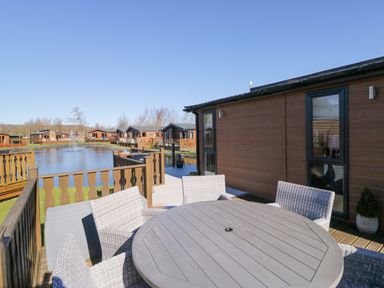 An outdoor deck area with a round table and wicker chairs next to a pond with wooden cabins in the background at Coniston View Lodge in Warton near Carnforth