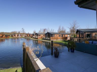 A view of a pond with wooden cottages along the shore and a patio with outdoor furniture at Coniston View Lodge in Warton near Carnforth