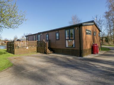 A brown wooden lodge with a deck and gas cylinders on the side in an outdoor setting at Coniston View Lodge in Warton near Carnforth