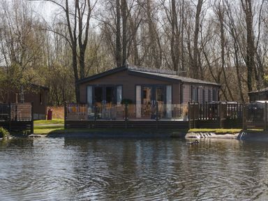 A wooden lodge with large windows and a fenced deck by a pond with ducks at Coniston View Lodge in Warton near Carnforth