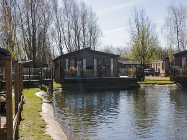 A lake in front of wooden lodges with glass doors and fenced decks at Coniston View Lodge in Warton near Carnforth