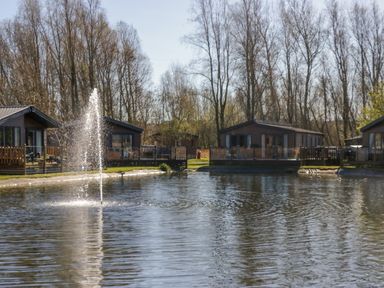 A pond with a fountain in front of several wooden lodges and trees at Coniston View Lodge in Warton near Carnforth