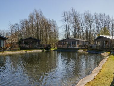 A pond surrounded by grass and four wooden lodges with glass railings and trees in the background at Coniston View Lodge in Warton near Carnforth