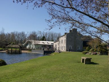 A stone building and a modern building beside a pond with a wooden gazebo and a stone bench on the grass at Coniston View Lodge in Warton near Carnforth