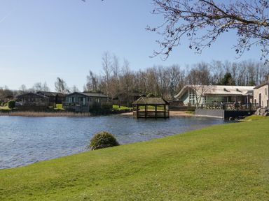 A lakeside scene with a grassy bank a gazebo over the water and buildings in the background at Coniston View Lodge in Warton near Carnforth