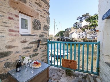 A small outdoor area with a table holding a bottle of prosecco two glasses and a plate of scones beside a blue railing overlooking boats and houses at Harbour View in Polperro