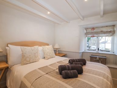 A bedroom with a double bed with pillows and folded towels on it a window with a floral blind and small tables with lamps at Harbour View in Polperro