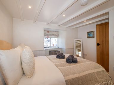 A bedroom with a bed rolled towels on the bed a window with floral blind a wicker basket a standing mirror a wooden door and a framed painting at Harbour View in Polperro