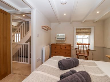 A bedroom with a bed with folded towels a wooden dresser with a mirror a wooden chair an open door showing a staircase at Harbour View in Polperro