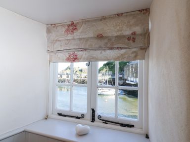A window with floral curtains overlooking boats on water at Harbour View in Polperro