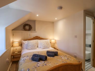 A bedroom with a double bed with floral bedding and three rolled towels two bedside tables with lamps and an open door leading to a bathroom at Harbour View in Polperro