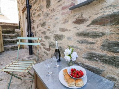 A metal chair next to a table with a bottle of wine two glasses a vase with white flowers and a plate of scones and strawberries against a stone wall at Harbour View in Polperro